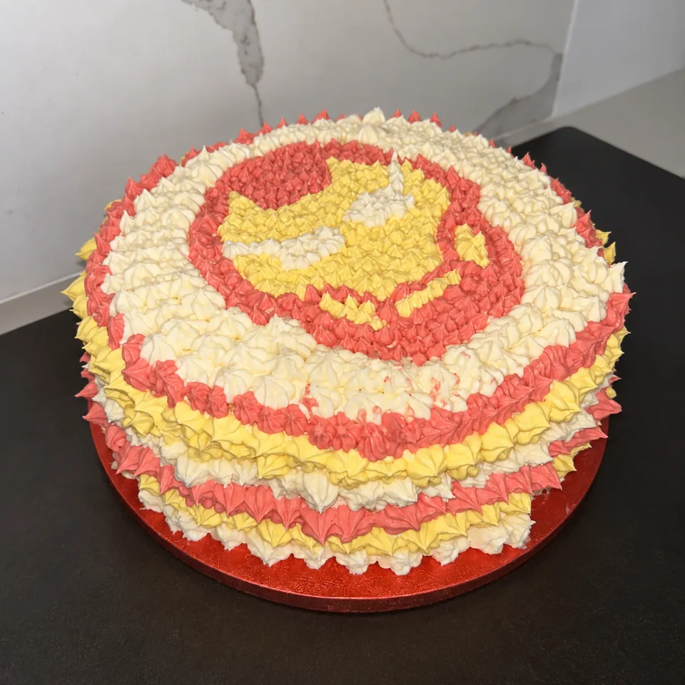 A round celebration cake decorated with yellow and red piped icing and a superhero design