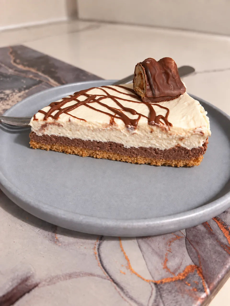 A slice of layered cheesecake with chocolate drizzle and a chocolate topping on a grey plate