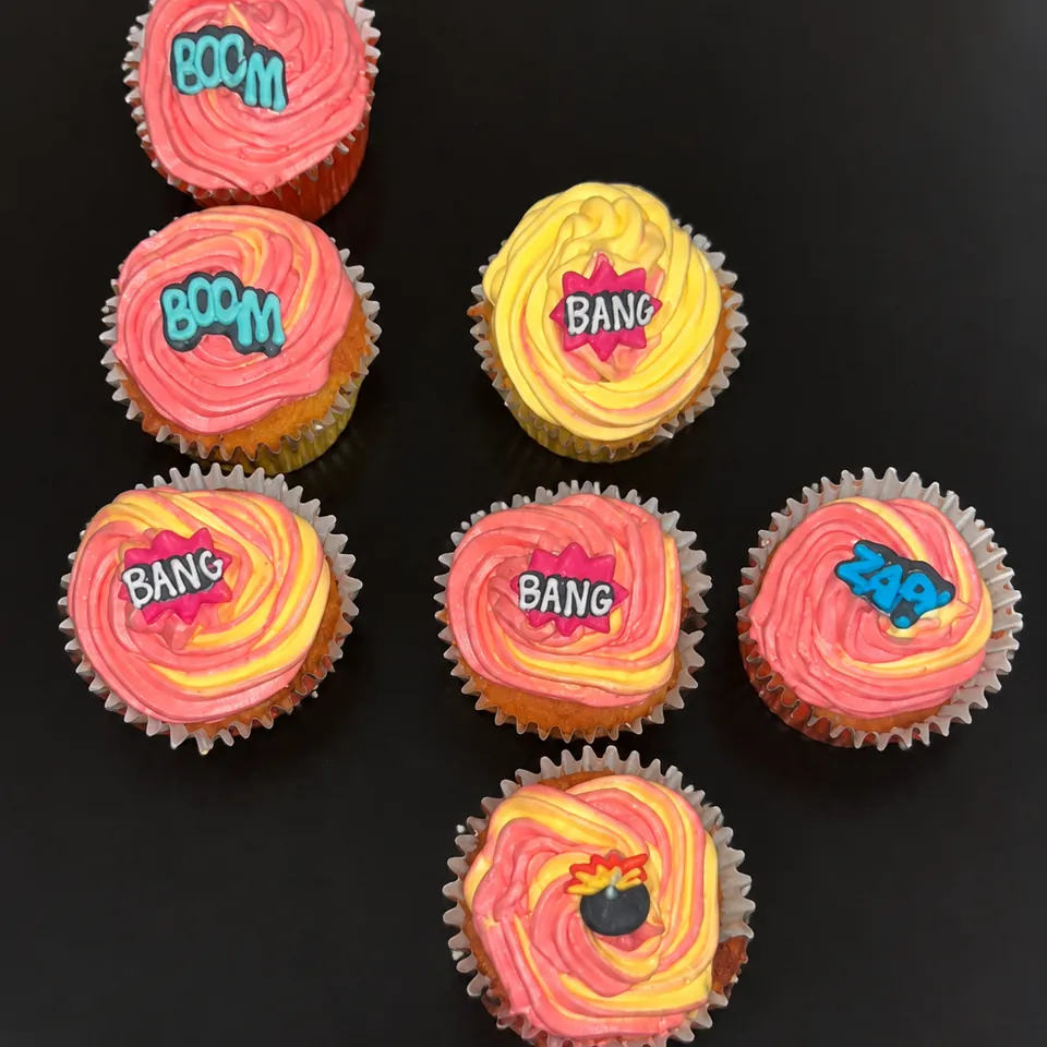 Homemade cupcakes with pink and yellow swirled icing and colourful comic-style toppers