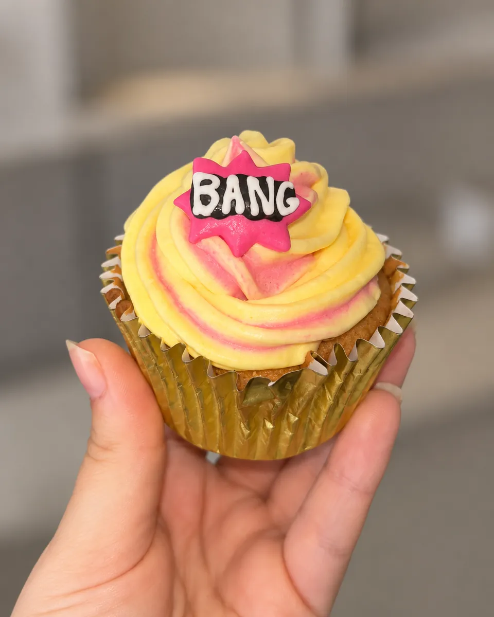 A hand holding a cupcake with yellow and pink swirled icing and a BANG topper in a gold case