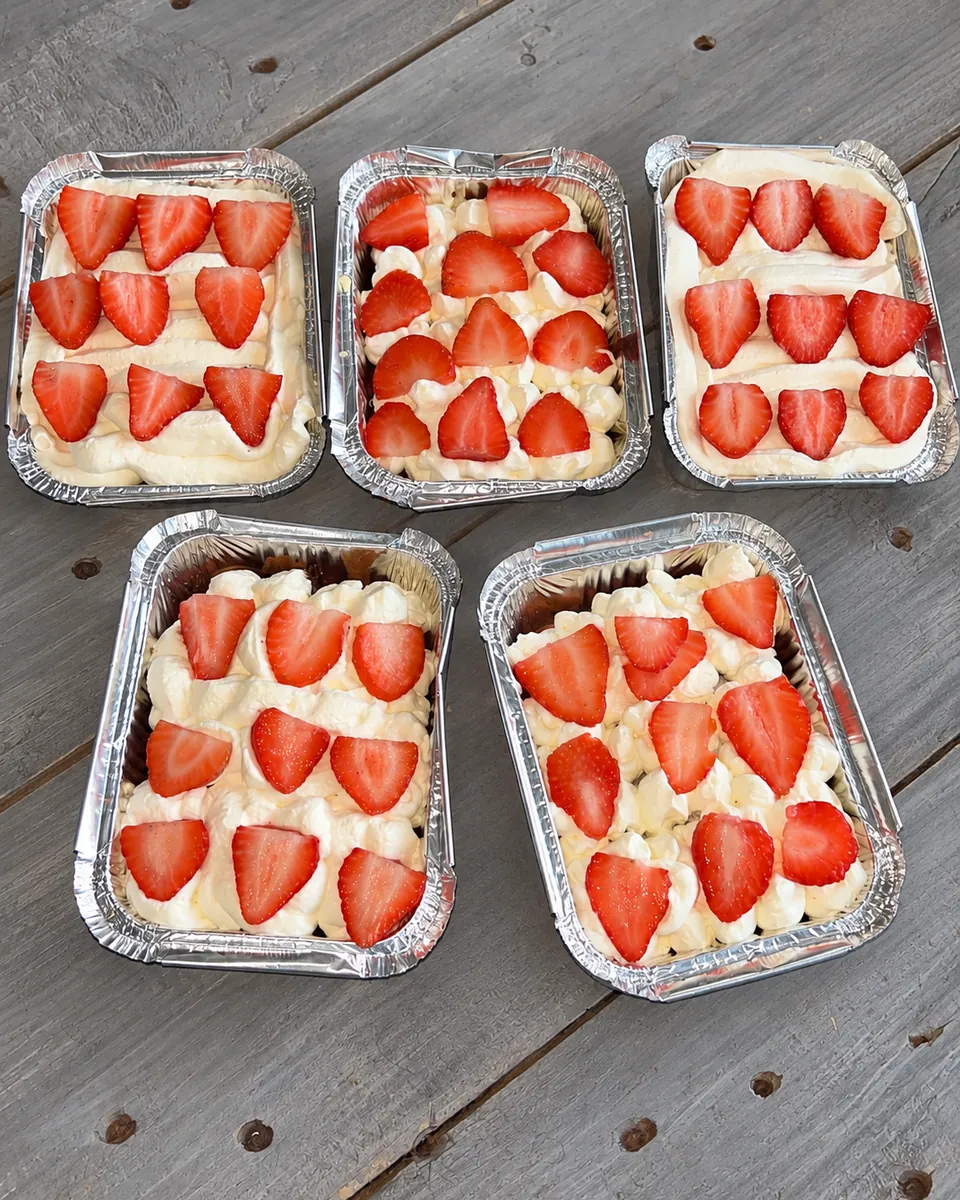 Five homemade bakes topped with cream and sliced strawberries, in foil trays on a wooden table